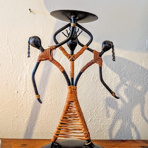 May include: A black metal candle holder with a circular base and a top for a candle. The holder features three stylized figures with woven rattan-like material and outstretched arms. The figures have black heads and arms, with gold accents.