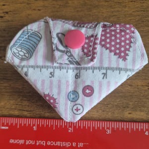 May include: A diamond-shaped fabric pouch featuring a pink and white striped pattern with sewing-themed illustrations of spools, scissors, and buttons. The pouch has a pink snap closure and is positioned above a red ruler measuring 10 cm.
