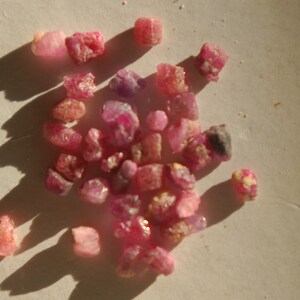 May include: A collection of small, pink and red ruby crystals. The crystals are rough and have a natural, unpolished look.