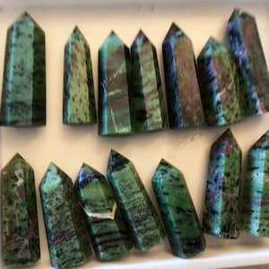 Puede incluir: Una colección de 14 puntas de piedras preciosas verdes, negras y rosas en una caja blanca. Las puntas son todas de tamaño y forma similares, y están dispuestas en una fila ordenada.