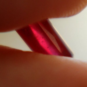 Könnte beinhalten: Nahaufnahme eines tief roten Edelsteins, möglicherweise ein Rubin, der zwischen zwei Fingern gehalten wird.