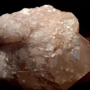 May include: A raw, natural quartz crystal cluster. The translucent, pale peach-colored crystal has a rough, textured surface with multiple facets and a crystalline structure. The crystal is set against a dark background, highlighting its natural beauty.