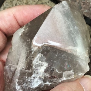 May include: A large, clear quartz crystal with a smoky brown hue. The crystal has a natural, rough surface and a pointed tip. The crystal is held in a hand, showing its size and shape.