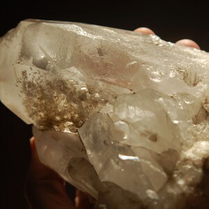 May include: A large, clear quartz crystal cluster with a rough, natural texture. The crystal is translucent and has a slight brown tint. The cluster is composed of multiple points and facets.