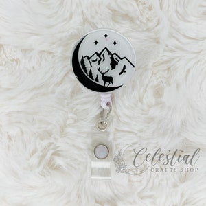 Peut inclure: Un porte-badge noir et blanc avec un motif de lune, de montagnes, d'étoiles et de cerf. Le porte-badge est attaché à un cordon rétractable blanc et à un porte-badge transparent. Le texte "Celestial Crafts Shop" est visible dans le coin inférieur droit de l'image.