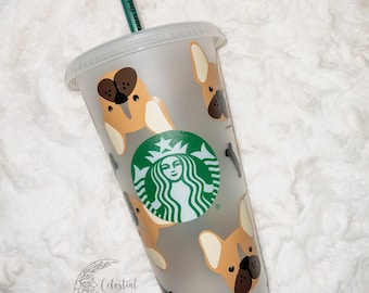 May include: A clear plastic reusable cup with a green Starbucks logo and a pattern of brown and white French bulldog faces. The cup has a green straw and a white background.