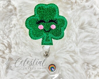 Puede incluir: Trébol verde con purpurina con una cara sonriente y mejillas rosadas, unido a un portagafete transparente con un diseño de arcoíris.  Celestial Crafts Shop está impreso en el portagafete.