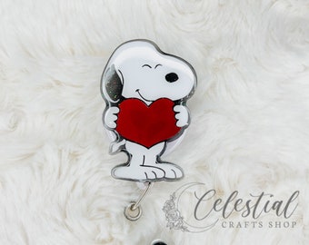 May include: A white and black cartoon dog holding a red heart, attached to a clear retractable badge holder with a black and green glitter button. "Celestial Crafts Shop" is printed on the bottom of the image.