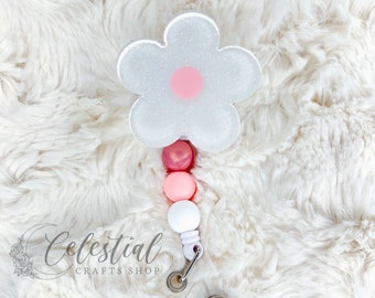 May include: A white glitter flower badge reel with a pink centre. The reel has a gradient of pink and white beads, a silver clip, and a clear plastic ID holder with a pink heart. The text "Celestial Crafts Shop" is visible.