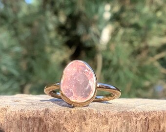 May include: A gold ring with a pink oval-shaped gemstone. The ring is sitting on a piece of wood.