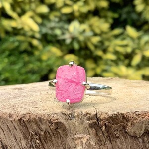 Peut inclure: Une bague en argent avec une pierre précieuse rose sertie sur quatre griffes. La bague est posée sur une surface en bois.