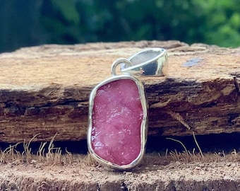 May include: A silver pendant featuring a pink, rough-cut gemstone. The pendant is set against a wooden background.