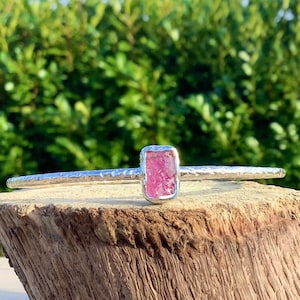 Op de afbeelding: Een zilveren armband met een roze, rechthoekige, ruwe edelsteen in het midden.