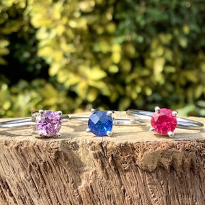 May include: Three silver rings with different coloured gemstones. The rings are on a piece of weathered wood. The gemstones are pink, blue, and red.