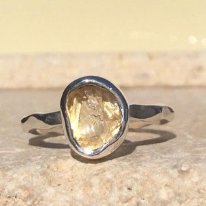 Puede incluir: Un anillo de plata con una piedra preciosa amarilla rugosa engastada en un engaste de bisel. El anillo tiene una textura martillada.