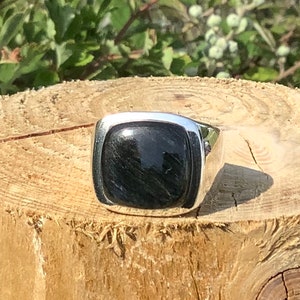 May include: A silver ring with a black square gemstone. The ring is sitting on a wooden surface.