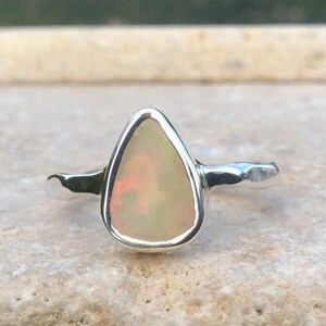 Peut inclure: Une bague en argent avec une pierre d'opale en forme de larme. L'opale est de couleur blanc laiteux avec des nuances de rose et de vert.
