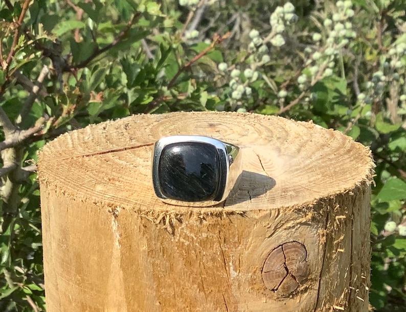 May include: A silver ring with a black square gemstone. The ring is sitting on a wooden surface.