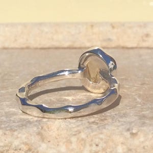 Puede incluir: Un anillo de plata con una banda texturizada y una piedra preciosa redonda, ligeramente ovalada. La piedra preciosa es de color amarillo claro.