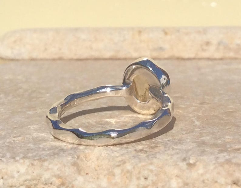 Puede incluir: Un anillo de plata con una banda texturizada y una piedra preciosa redonda, ligeramente ovalada. La piedra preciosa es de color amarillo claro.