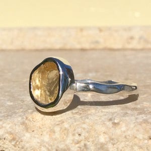 Puede incluir: Un anillo de plata con una piedra preciosa amarilla engastada en un engaste de bisel. La banda del anillo es texturizada y tiene un dise&ntilde;o ondulado.