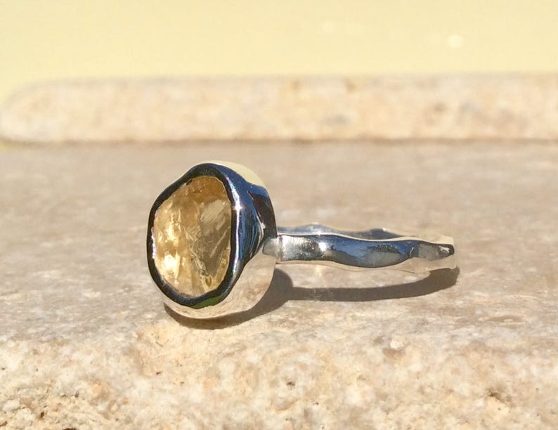 Puede incluir: Un anillo de plata con una piedra preciosa amarilla engastada en un engaste de bisel. La banda del anillo es texturizada y tiene un dise&ntilde;o ondulado.