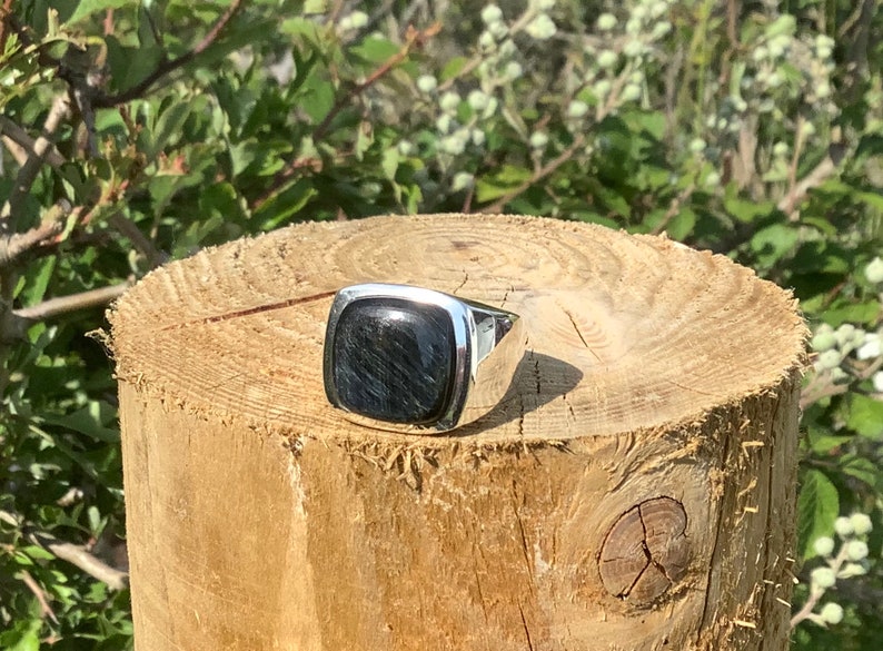 May include: A silver ring with a square black gemstone. The ring is sitting on a wooden surface.