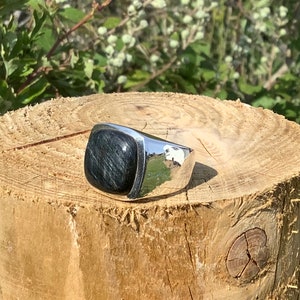 May include: A silver ring with a square-shaped black gemstone. The ring is sitting on a wooden surface.