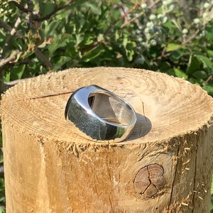 May include: A silver ring with a smooth, rounded, oval-shaped top. The ring is sitting on a wooden surface.