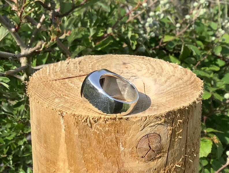 May include: A silver ring with a smooth, rounded, oval-shaped top. The ring is sitting on a wooden surface.