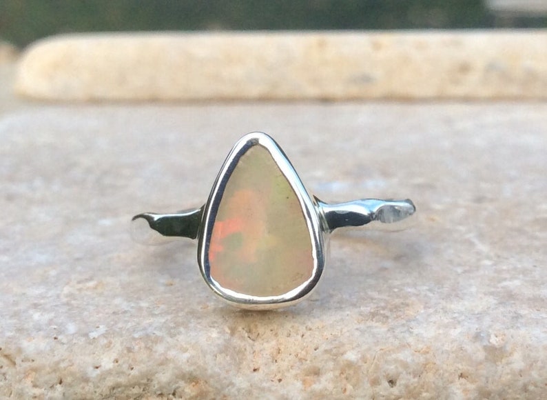 Peut inclure: Une bague en argent avec une pierre d'opale en forme de larme. L'opale est de couleur blanc laiteux avec des nuances de rose et de vert.