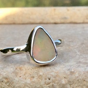 Peut inclure: Une bague en argent avec une pierre d'opale en forme de larme. L'opale est de couleur blanche avec des nuances de rose et de vert.