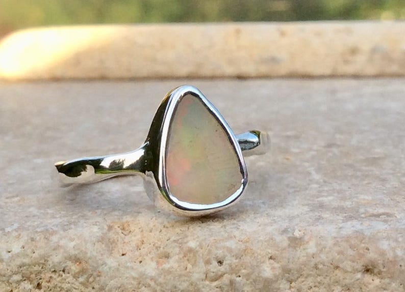 Peut inclure: Une bague en argent avec une pierre d'opale en forme de larme. L'opale est de couleur blanche avec des nuances de rose et de vert.