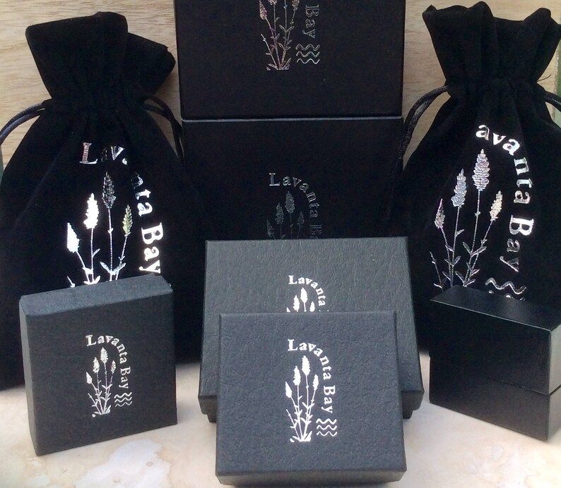 Peut inclure: Des pochettes en velours noir avec des lettres argent&eacute;es qui lisent "Lavanta Bay" et un motif floral argent&eacute;. Les pochettes sont pos&eacute;es sur des bo&icirc;tes noires avec les m&ecirc;mes lettres et le m&ecirc;me motif.