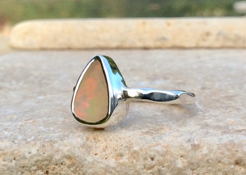 Peut inclure: Une bague en argent avec une pierre d'opale en forme de larme. L'opale est de couleur rose p&acirc;le avec un l&eacute;ger &eacute;clat iridescent.