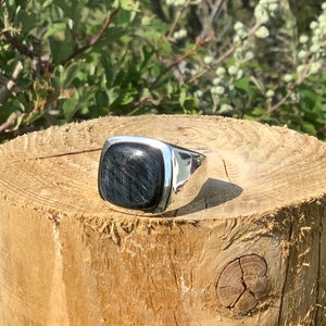 May include: A silver ring with a square-shaped blue gemstone. The ring is sitting on a wooden surface.