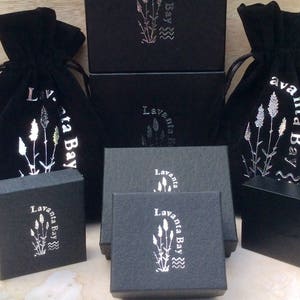 Puede incluir: Bolsas de terciopelo negro con letras plateadas que dicen "Lavanta Bay" y una imagen estilizada de flores de lavanda. Las bolsas se muestran sobre cajas negras con la misma marca.