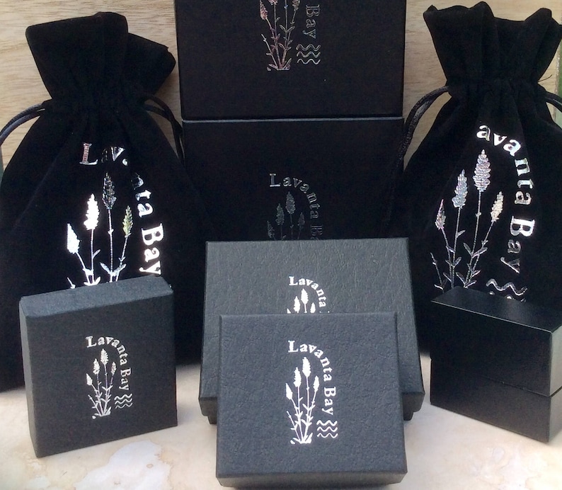 Puede incluir: Bolsas de terciopelo negro con letras plateadas que dicen "Lavanta Bay" y una imagen estilizada de flores de lavanda. Las bolsas se muestran sobre cajas negras con la misma marca.