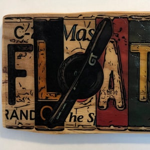 May include: A wooden sign with the word "FLAT" spelled out in colourful letters. The letters are painted in shades of yellow, red, blue, and green. A black wrench is positioned between the letters "L" and "A".