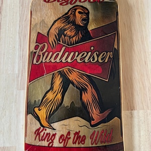 Könnte beinhalten: Ein Holzausschnitt einer Bierdose mit einer Bigfoot-Illustration. Die Dose ist rot, weiß und braun mit dem Text "Bigfoot", "Budweiser" und "King of the Wild".