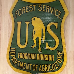 Puede incluir: Un letrero de madera amarillo y verde con el texto "FOREST SERVICE US FROGMAN DIVISION DEPARTMENT OF AGRICULTURE". El letrero presenta una rana de dibujos animados con un casco militar.