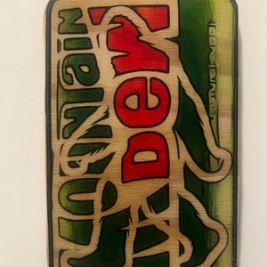 May include: Wooden sign in the shape of a soda can, featuring the words "BIGFOOT" and "MOUNTAIN DEW" in red and green. The design has a rustic, carved appearance, with a Bigfoot silhouette incorporated into the lettering.