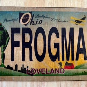 May include: A vintage-style sign with a green frog illustration, the text "FROGMAN" in black, and the words "Beautiful Ohio Birthplace of Aviation LOVELAND" in a stylized font.