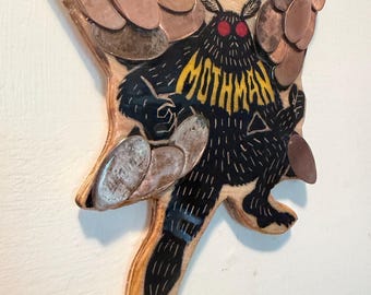 May include: A wooden wall hanging shaped like a moth, featuring a black illustration of the Mothman with red eyes and yellow text reading MOTHMAN. The wings are decorated with overlapping copper-colored metal coins or discs.