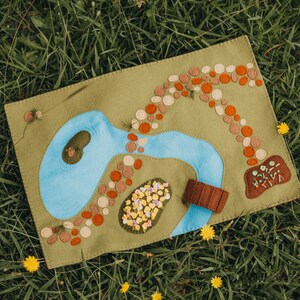 May include: A felt play mat depicting a landscape with a blue river, brown bridge, and colourful details. The mat includes a path of orange and tan circles, and a patch of embroidered flowers. The mat is set on green grass.