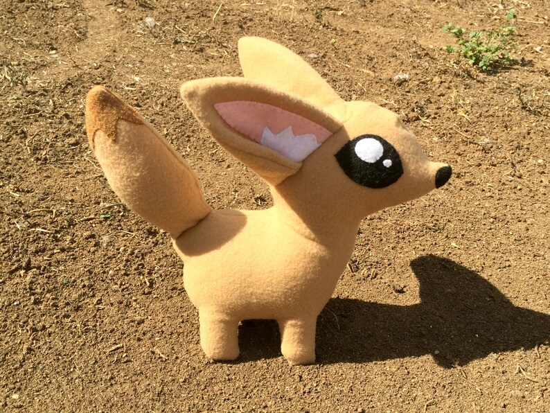 Peut inclure: Peluche repr&eacute;sentant un fennec, de couleur beige clair, avec de grandes oreilles aux int&eacute;rieurs roses et blancs, une longue queue et de grands yeux noirs et blancs. Le renard est pos&eacute; sur quatre petites pattes.