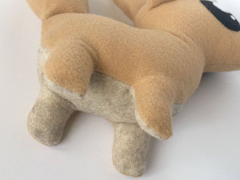 Peut inclure: Une peluche en forme de chien, principalement beige avec des accents beige clair sur les pattes et le ventre. L'&oelig;il du chien est noir et blanc. Le jouet est fait d'une mati&egrave;re douce semblable &agrave; du feutre.