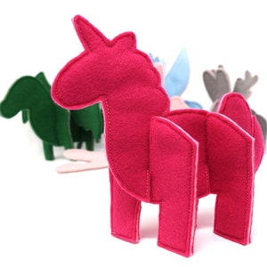 May include: A felt unicorn toy in bright pink, with a white stitched outline. The unicorn has a horn and is standing upright. Other felt animal toys in green, blue, and gray are visible in the background. The toys are designed for children.