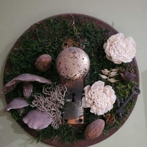May include: A round, rustic wall hanging featuring a vintage door knob and keyhole. The design incorporates green moss, white flowers, mushrooms, nuts, and a decorative key. The arrangement is set on a dark brown, circular base.