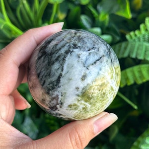 May include: A green, white, and black sphere made of polished stone. The sphere is being held in a hand.